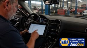 A mechanic troubleshooting a car's climate control system in Gregg Smith Automotive, with a rain-speckled windshield offering a view of the workshop and diagnostic equipment.