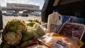 A close-up, dramatic shot inside the dark, intensely hot rear cargo area of an SUV showing a grocery haul rapidly spoiling, including wilted lettuce, sweating milk cartons, and thawed meat packages, with the unshaded gravel parking lot of the Market of Marion visible in the background under the Florida sun.